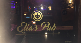 Blick in den Pub durch die geschlossene Glastür auf der der goldene Schriftzur Elias Pub sowie das Logo ein schwarzes Pik in goldenem Rund prangt